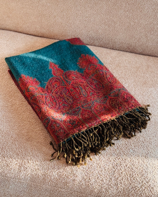 Coperta in lana jacquard verde petrolio con bordo paisley rosso, piegata sul divano