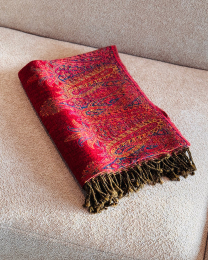 Coperta in lana jacquard rossa con paisley oro e blu piegata sul divano, con frange