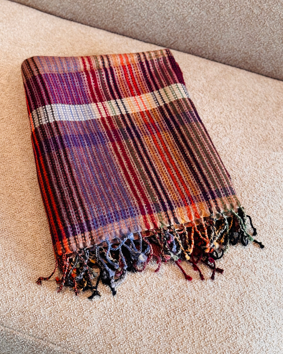 Plaid in lana merino a quadri viola, rosso e arancio piegato sul divano, con frange