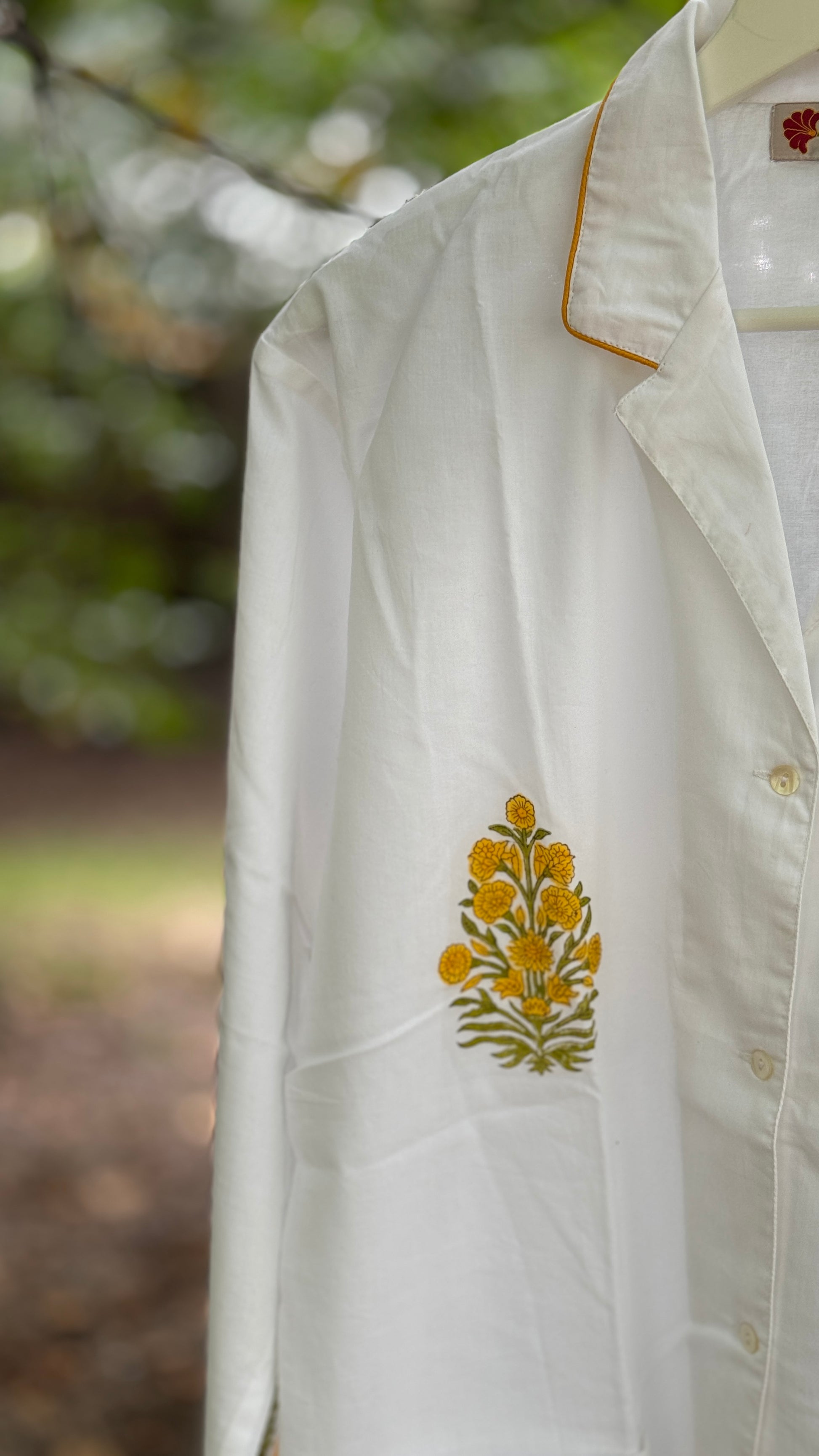 Pigiama due pezzi in cotone voile bianco con stampe floreali Buti ocra fatte a mano, appeso tra rami verdi in un contesto naturale.