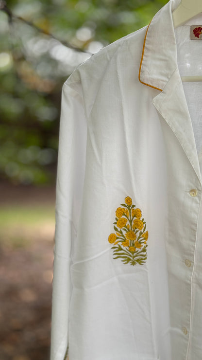 Pigiama due pezzi in cotone voile bianco con stampe floreali Buti ocra fatte a mano, appeso tra rami verdi in un contesto naturale.