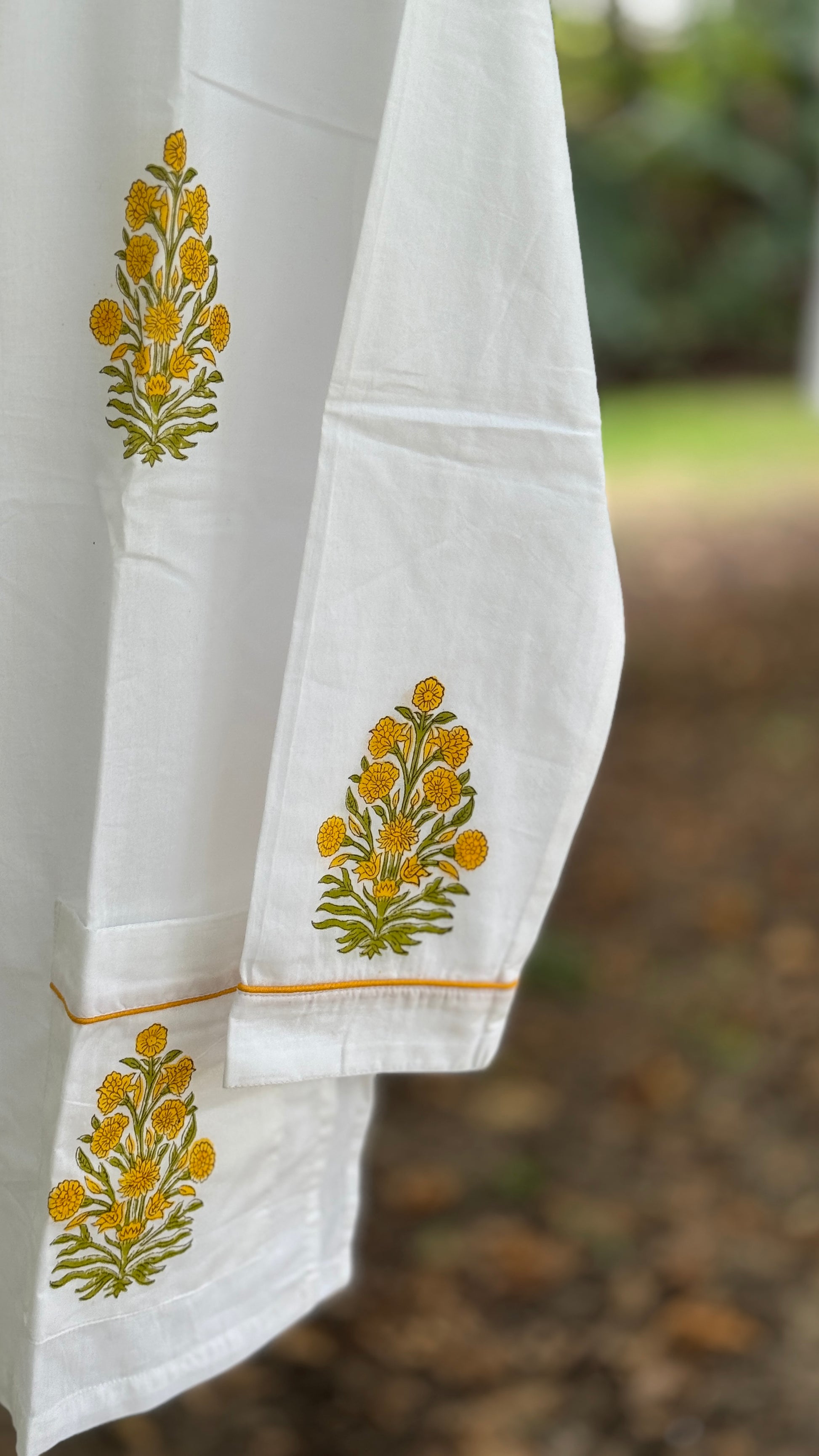 Pigiama due pezzi in cotone voile bianco con stampe floreali Buti ocra fatte a mano, appeso tra rami verdi in un contesto naturale.