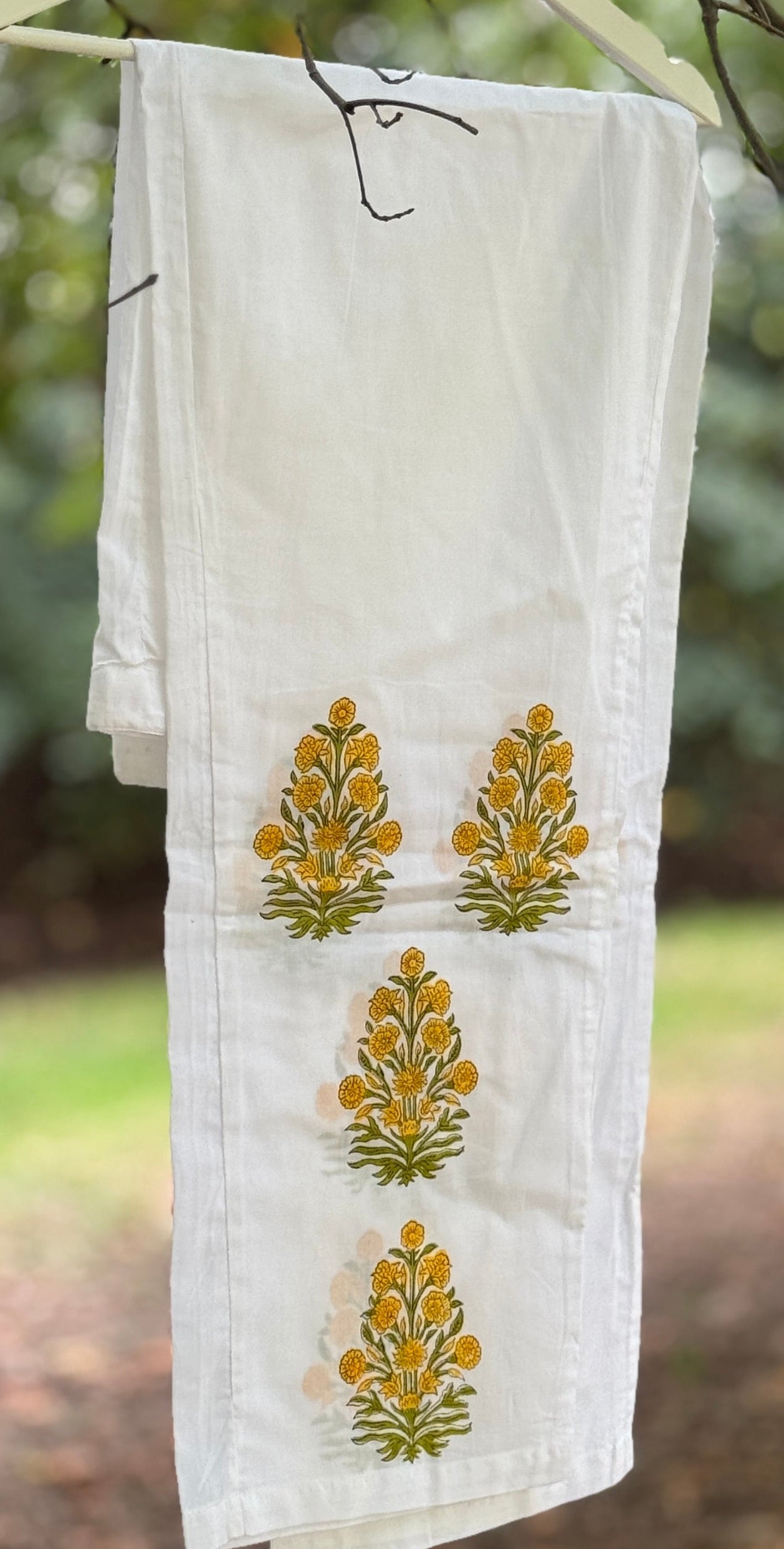 Pigiama due pezzi in cotone voile bianco con stampe floreali Buti ocra fatte a mano, appeso tra rami verdi in un contesto naturale.